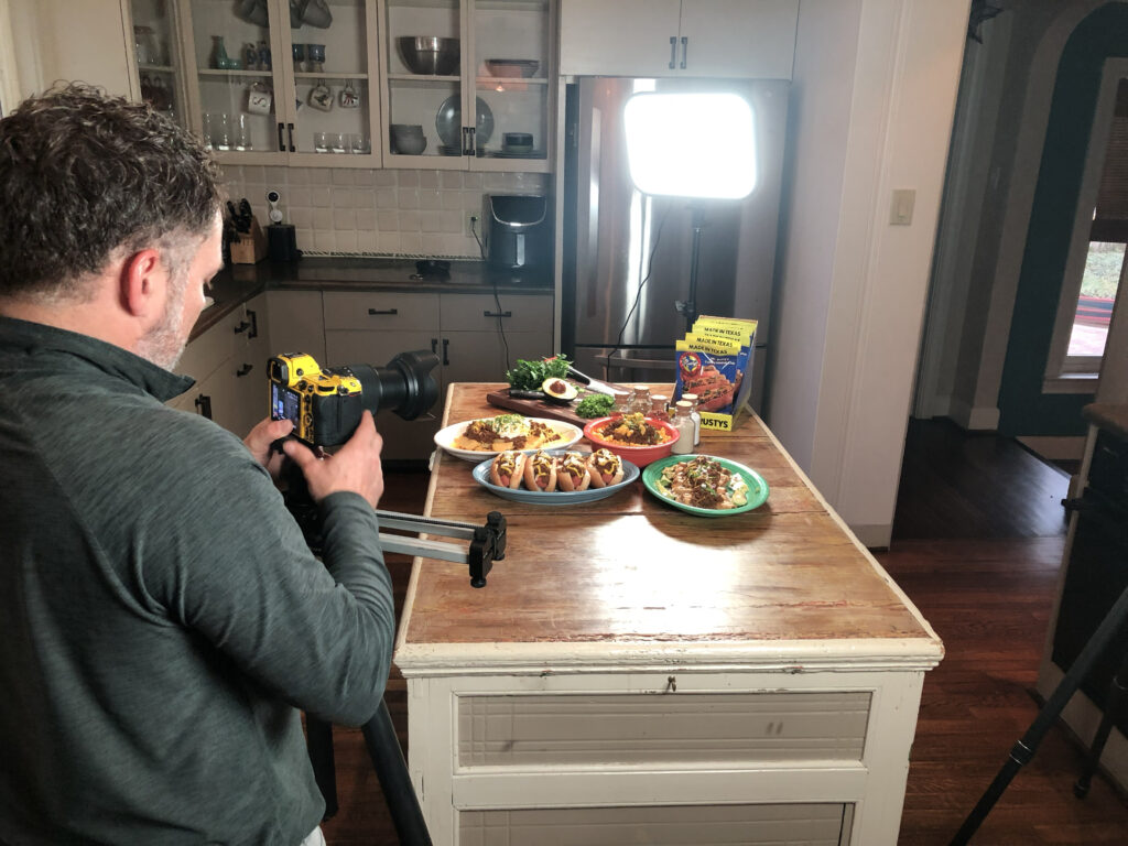 Photographer Patrick Bertolino preparing a food photography setup with tripod, soft lighting, and yellow-accented Nikon camera.